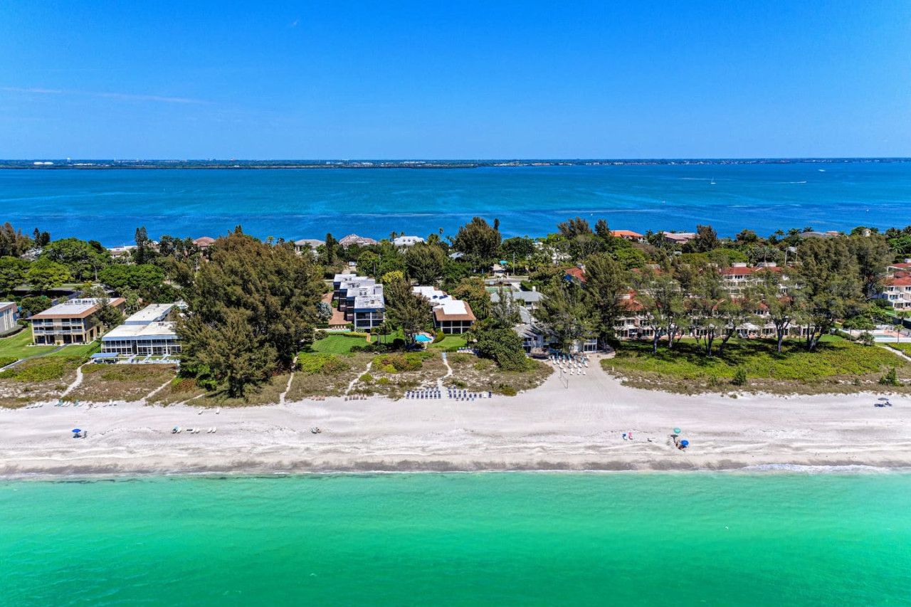 An aerial view of a coastal area with buildings, a beach, and clear blue waters.