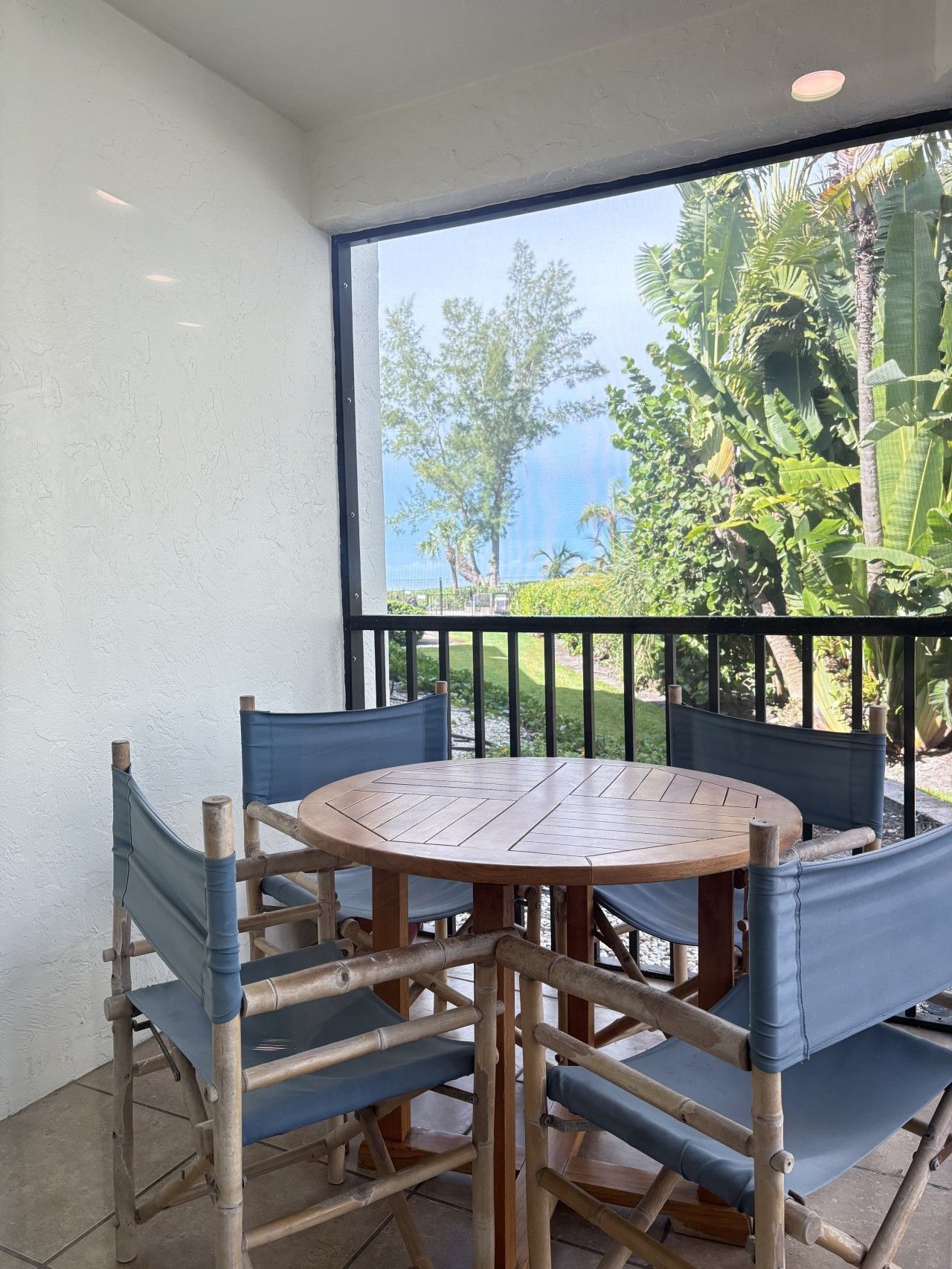 A round wooden table with six chairs, a balcony with a view of greenery and a building, and a white wall.
