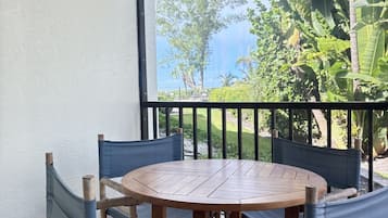 A round wooden table with six chairs, a balcony with a view of greenery and a building, and a white wall.