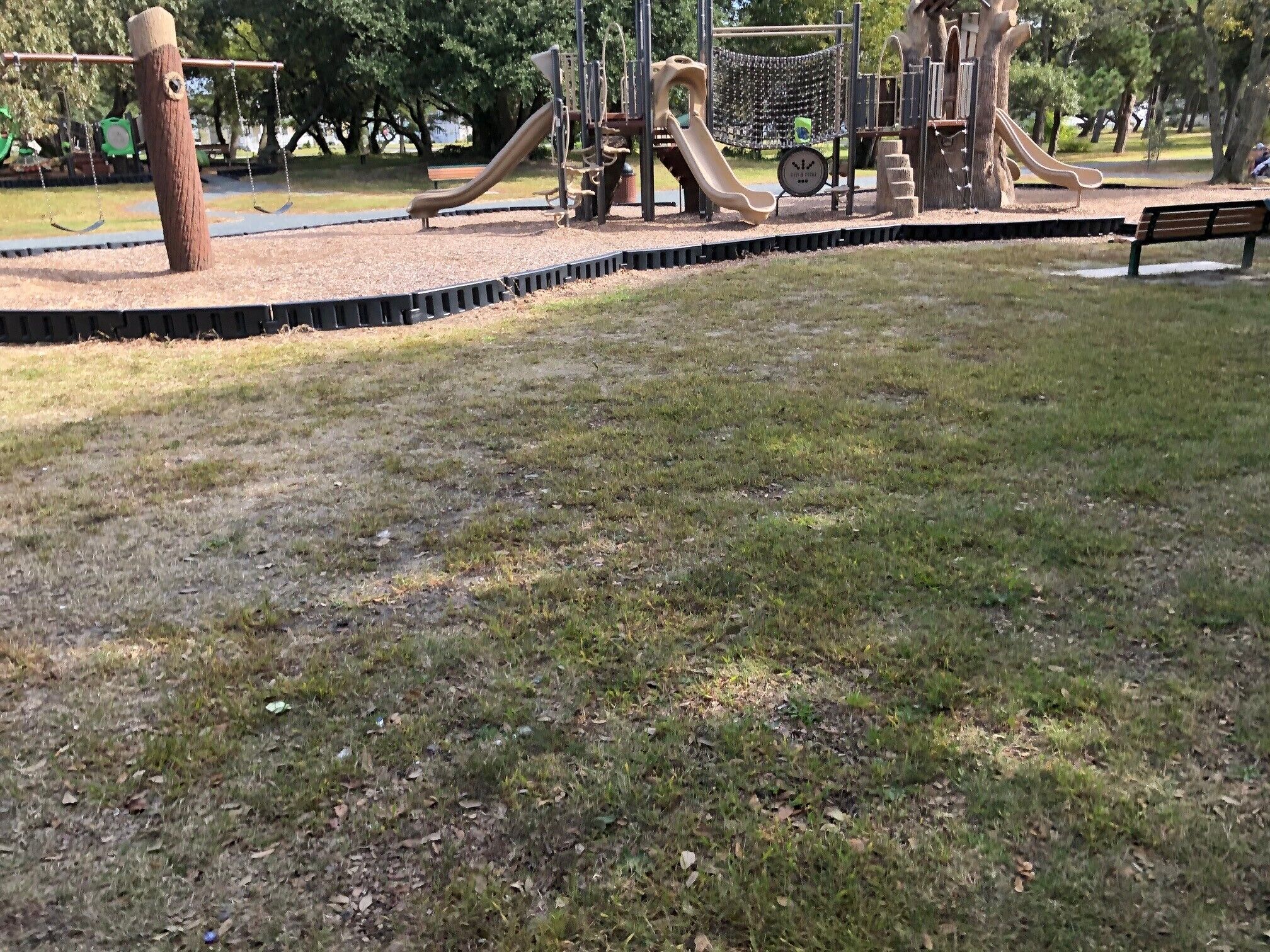 A playground with slides and climbing structures, surrounded by a grassy area and trees.