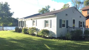 A single-story house with a porch, a small deck, and a grassy yard. - Port Bay Waterfront Cottage — Your Private Lakeside Escape (Wolcott)