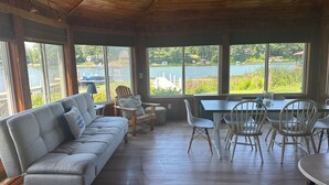 A room with a wooden ceiling, large windows, a grey sofa, a dining table with chairs, and a view of a lake. - Port Bay Waterfront Cottage — Your Private Lakeside Escape (Wolcott)