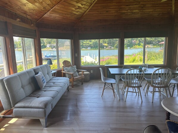 A room with a wooden ceiling, large windows, a grey sofa, a dining table with chairs, and a view of a lake.