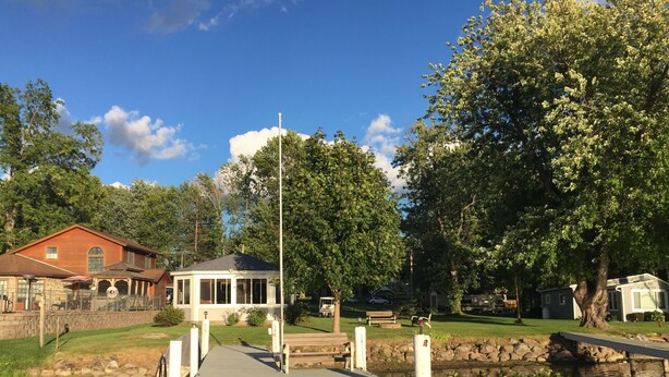 A wooden dock leading to a gazebo with a glass roof, surrounded by a pond with lily pads, and a red building in the background. - Port Bay Waterfront Cottage — Your Private Lakeside Escape (Wolcott)
