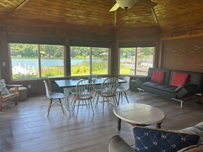 A spacious room with a wooden ceiling, large windows overlooking a lake, a dining table with chairs, a sofa with red cushions, and a small round table.