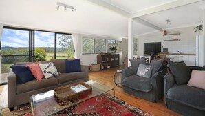 A spacious living room with a large window, a sofa, a coffee table, and a television. - Twin Oaks Cottage - Fitzroy Falls  (Fitzroy Falls)