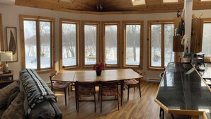 Interior acogedor de una cabaña con una mesa de madera, sillas, un sofá y ventanas amplias que ofrecen vistas a un paisaje nevado.