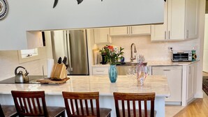A modern kitchen with a central island, wooden chairs, a stainless steel refrigerator, and a decorative fish wall art piece. - Ditch Plains - Walk to Ocean Beach - New Kitchen, Bath, & Bedrooms (Montauk)