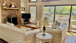A living room with white sofas, a TV, a fireplace, and a view of the outdoors. - Ditch Plains - Walk to Ocean Beach - New Kitchen, Bath, & Bedrooms (Montauk)