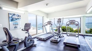 Ein Fitnessraum mit Cardiogeräten, einem Rudergerät und einem großen Fenster mit Meerblick.