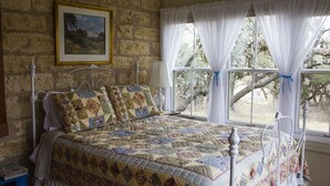 A bedroom with a stone wall, a bed with a quilt, a nightstand, and a window with curtains.