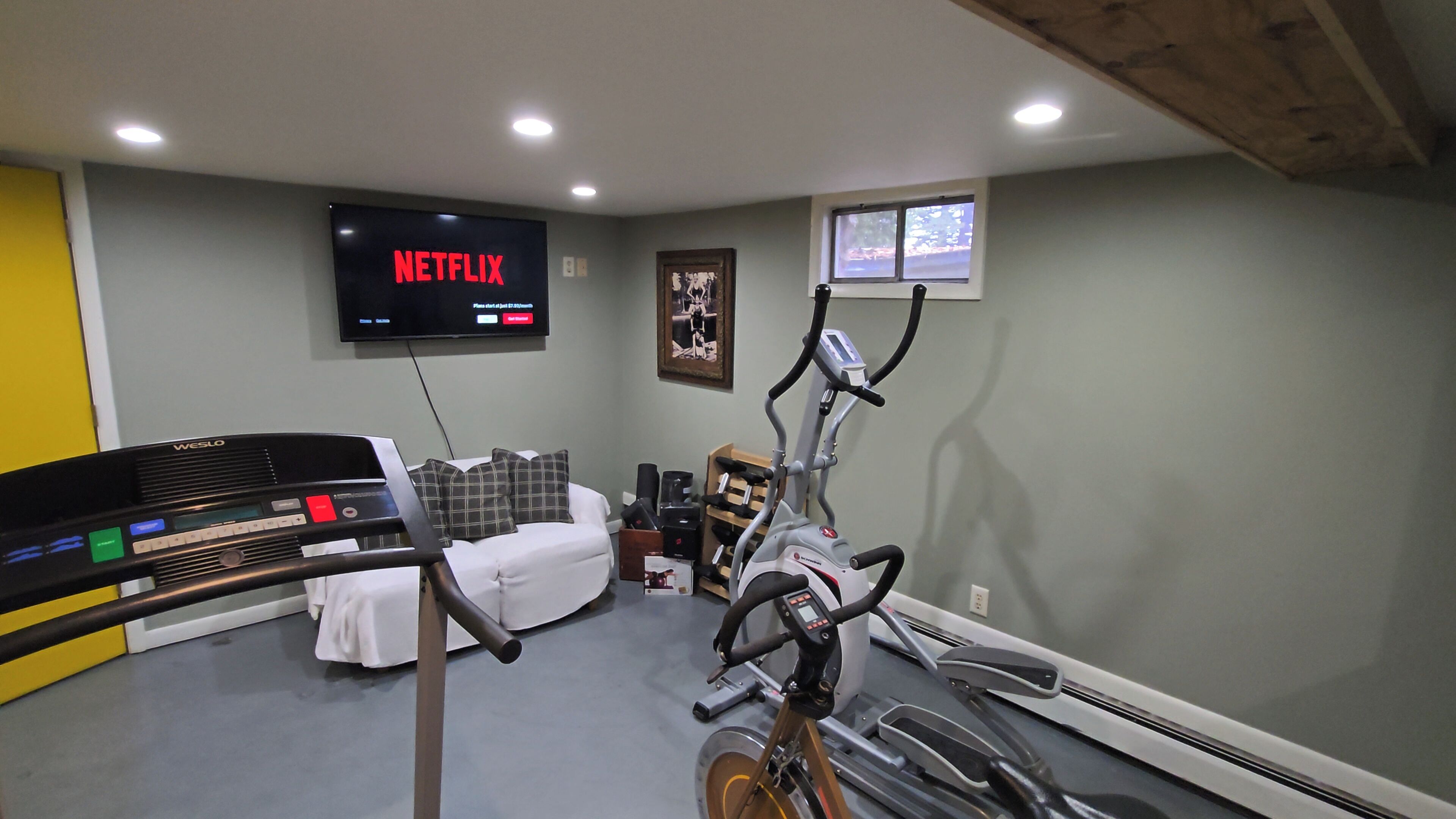 Et hjemmegymnasium med løbebånd, motionscykel og crosstrainer. Et tv på væggen viser 'NETFLIX'.