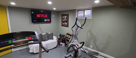 Et hjemmegymnasium med løbebånd, motionscykel og crosstrainer. Et tv på væggen viser 'NETFLIX'.