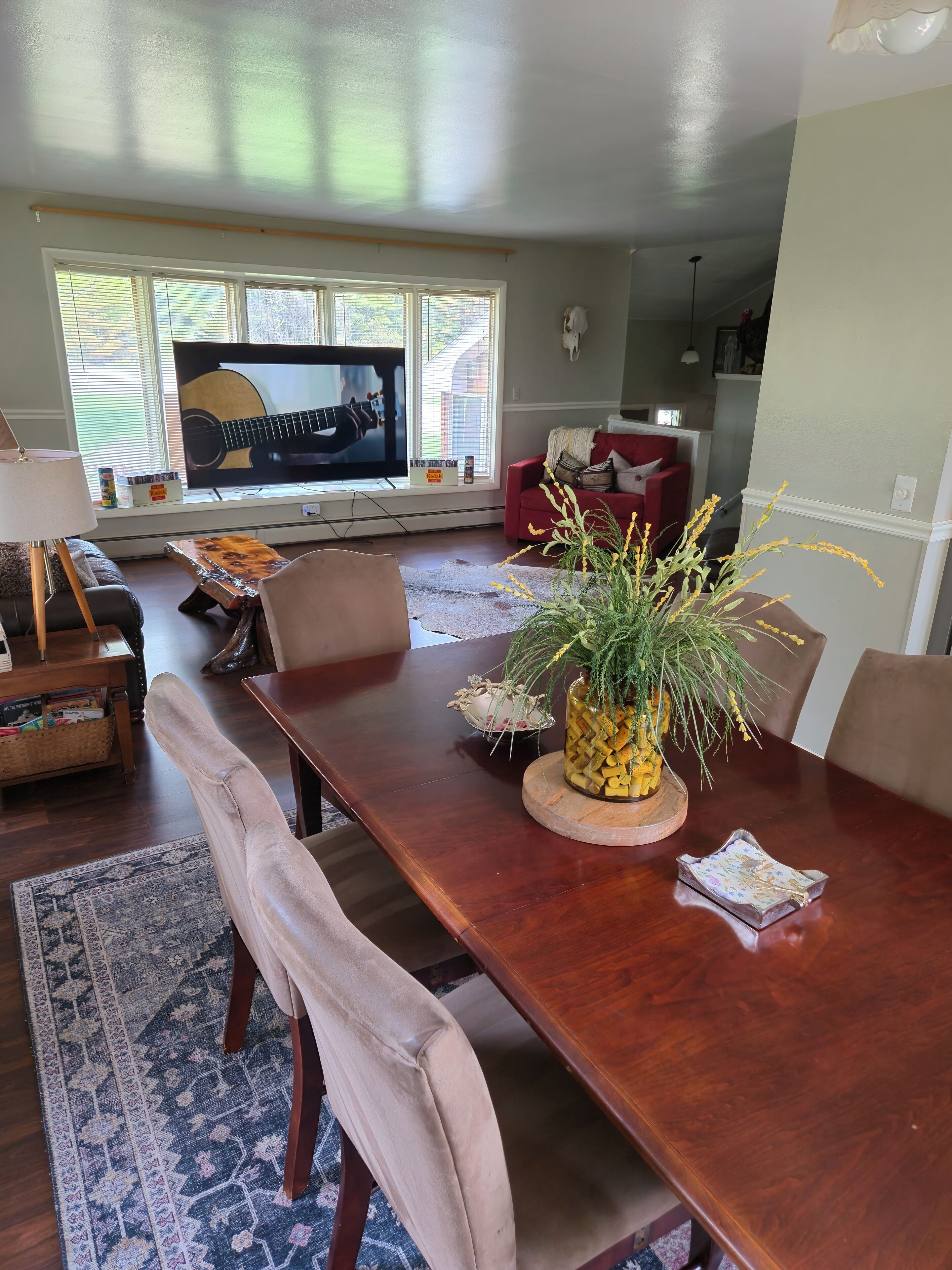 A dining area with a wooden table, chairs, a vase with plants, and a television displaying a guitar.