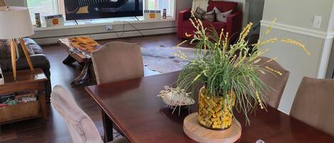 A dining area with a wooden table, chairs, a vase with plants, and a television displaying a guitar.