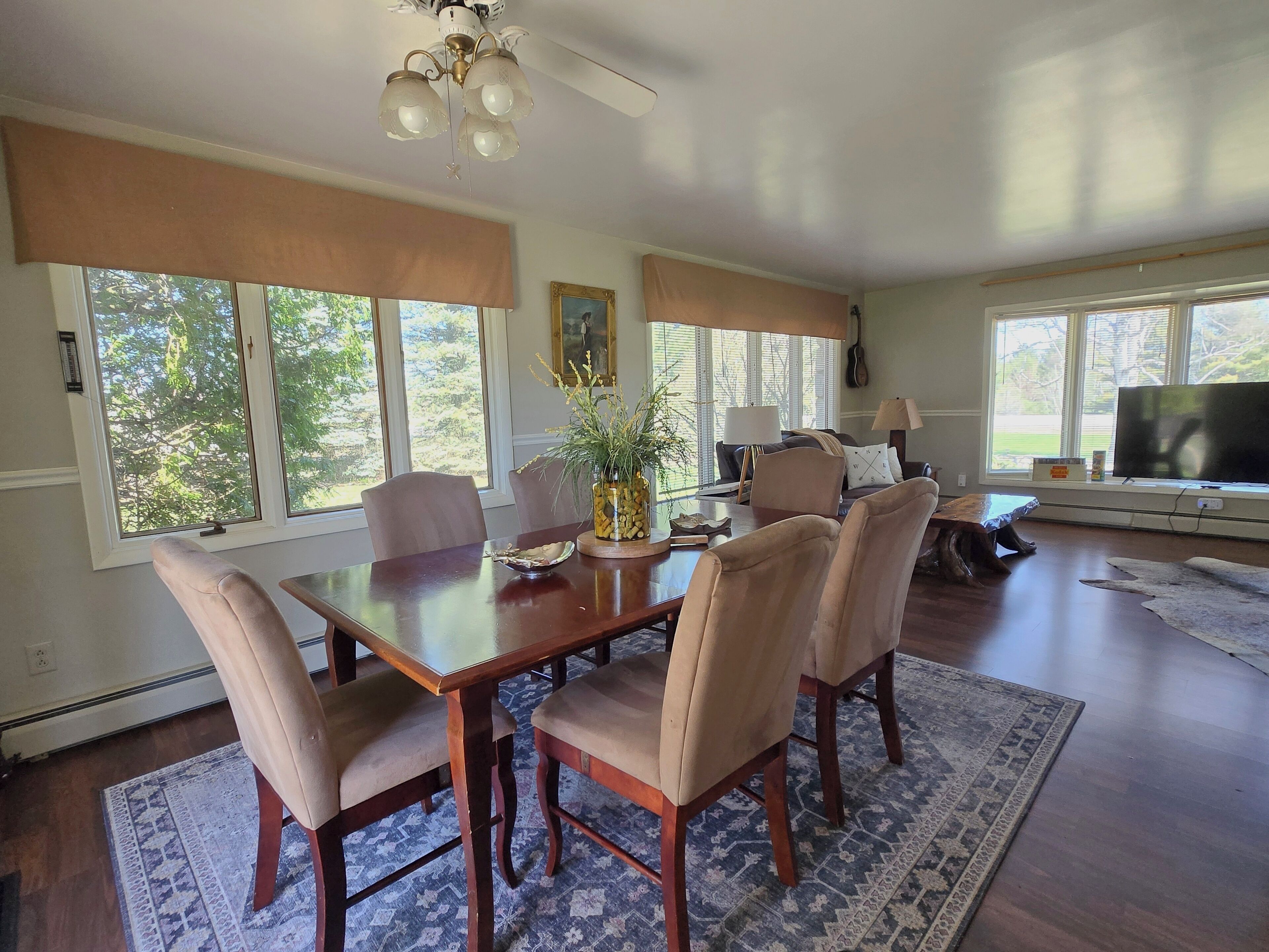 A dining area with a wooden table, beige upholstered chairs, a vase with flowers, and a large window with blinds.