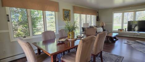A dining area with a wooden table, beige upholstered chairs, a vase with flowers, and a large window with blinds.