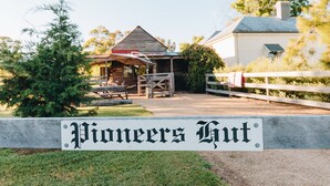 Ein rustikales Gebäude mit der Aufschrift 'Pioneers Hut', das einen Holzbau, eine überdachte Außen-Sitzgelegenheit und einen umzäunten Garten aufweist.