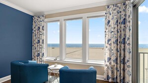 Ein Zimmer mit Blick auf den Strand, zwei blaue Sessel, ein kleiner Tisch und ein Fenster mit Vorhängen.