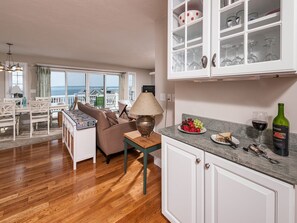 Uma sala de estar aconchegante com piso de madeira, um sofá, uma mesa de centro e um espaço de jantar com mesa e cadeiras. É visível um armário com portas de vidro, e há uma garrafa de vinho no balcão.
