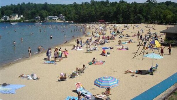 Una playa concurrida con gente tomando el sol y nadando, rodeada de árboles y un cielo despejado.