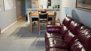 A living room with a leather couch, dining table, and chairs. There is a kitchen area in the background with a refrigerator and a window.