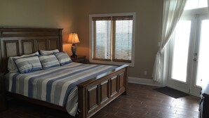 A bedroom with a large bed, wooden headboard, a nightstand with a lamp, a window with blinds, and a door with curtains. - "The Cut Pearl" - Stunning Private Beachfront/Oceanfront Villa On Little Exuma (Forbes Hill)