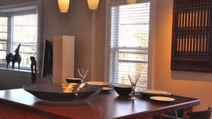 Un comedor con mesa de madera dispuesta para cuatro, ventanas amplias con persianas e iluminación colgante.