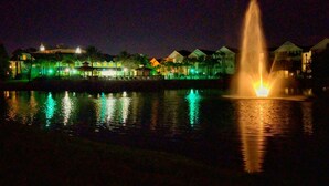 Une vue nocturne d’une zone riveraine, avec une fontaine, des bâtiments illuminés et des reflets sur l’eau.