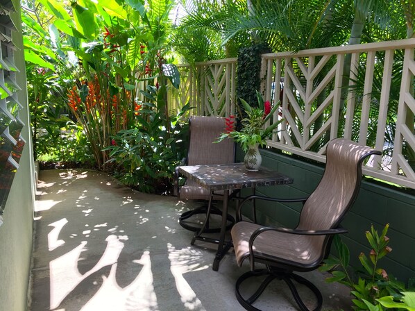 A patio area with a table and two chairs, surrounded by lush greenery and a wooden fence.