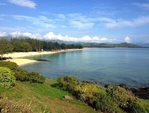 Paesaggio costiero con spiaggia, acqua limpida e montagne sullo sfondo.