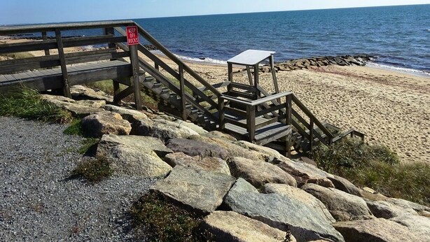 Un escalier en bois mène à une zone de plage où un panneau indique : « Défense de marcher sur les rochers ».