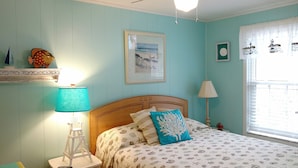 Quarto com cama, mesa de cabeceira, luminária, quadro emoldurado e janela com cortinas.