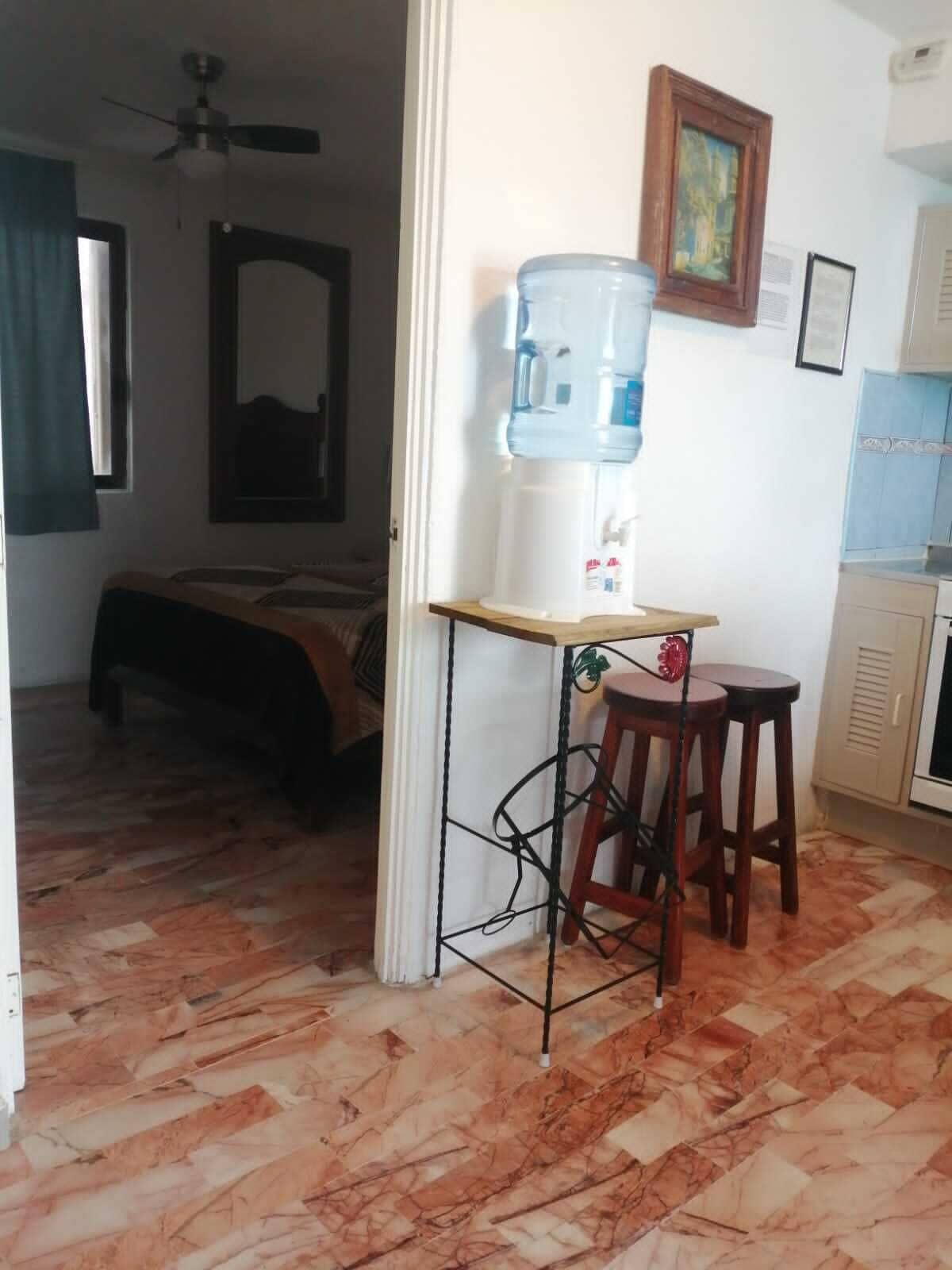 Une pièce comprenant un lit, un distributeur d’eau, un petit tabouret et un ventilateur de plafond.