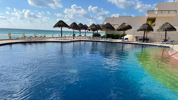 A clear blue pool with lounge chairs and thatched umbrellas, overlooking the ocean. -    Quality Cancun Condo Rental Within Your Vacation Budget?                      (Cancún)