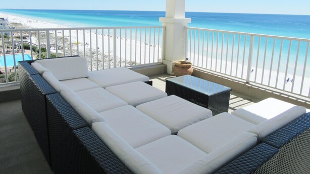 Een balkon met een witte, bekleedde bank en een kleine tafel, met uitzicht op een strand en een zwembad.