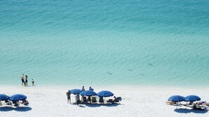 Een strand met helder turquoise water, verschillende mensen en blauwe parasols.