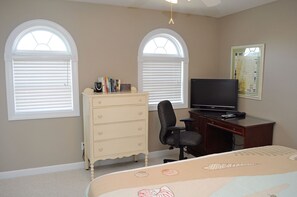 A bedroom with a bed, dresser, desk, chair, TV, and two windows with blinds.