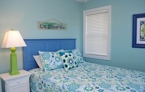 Quarto com cama, mesa de cabeceira, abajur, janela com persianas e um letreiro na parede.