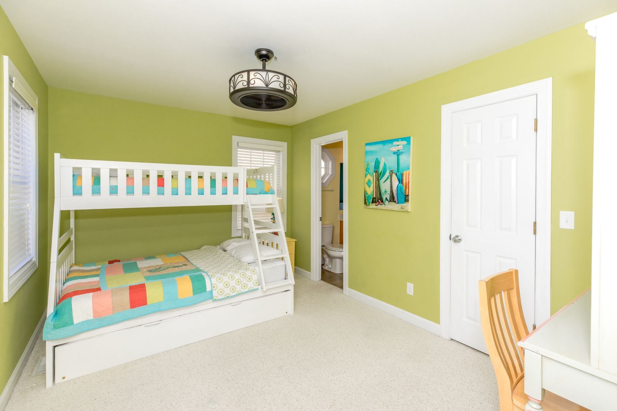 Una cama litera para niños con colcha de colores, un ventilador de techo, una puerta y un cuadro con tablas de surf en la pared.