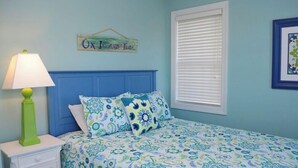 A bedroom with a blue headboard, a bed with patterned bedding, a bedside lamp, a window with blinds, and a sign on the wall. - C' Turtle - 5 BR Oceanfront Home - Elevator, Spectacular Views from Decks (Surf City)