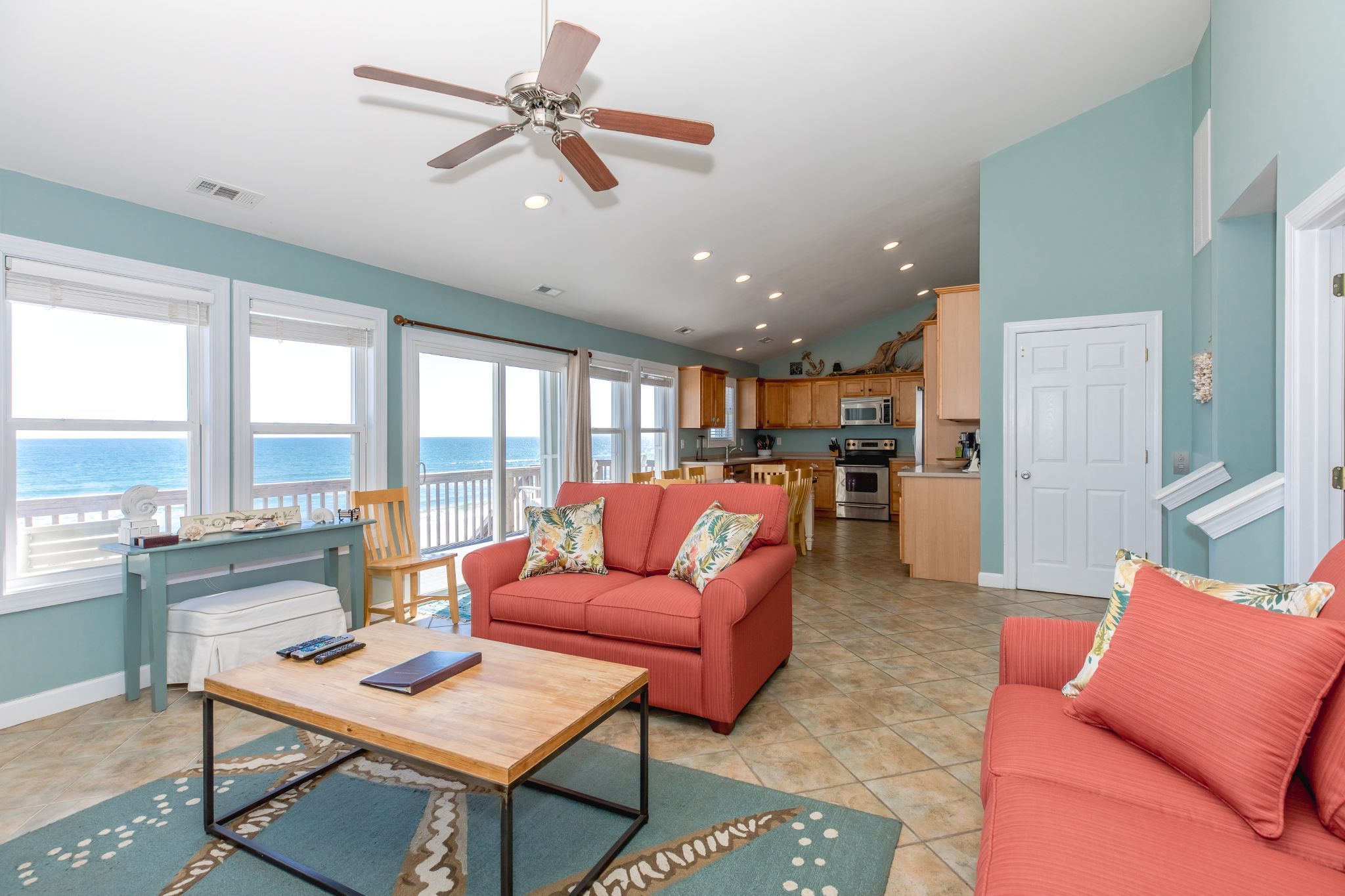 A spacious living area with a large window overlooking the ocean, a ceiling fan, and a kitchen with wooden cabinets.