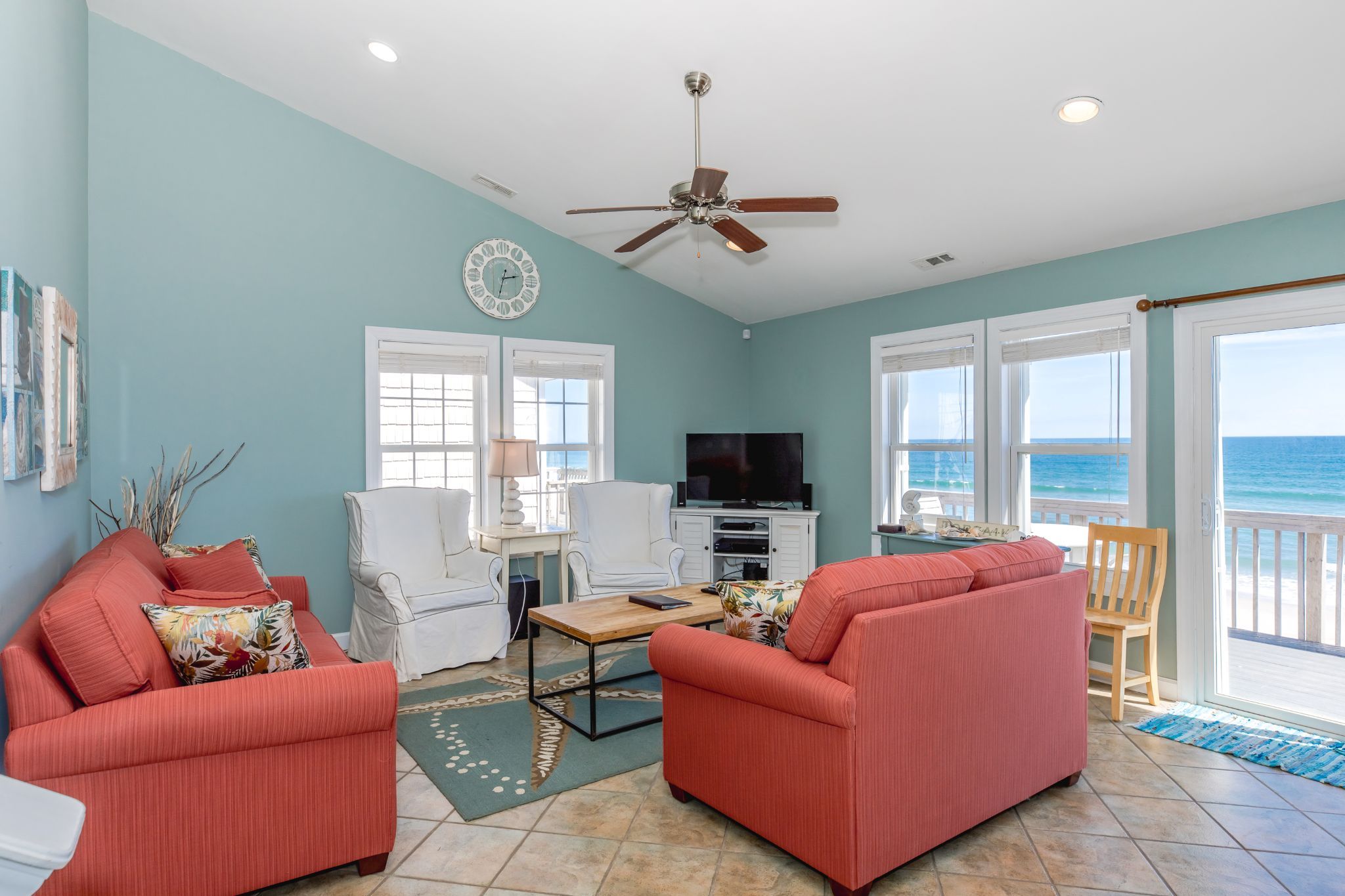 A living room with a ceiling fan, a television, and a view of the ocean through large windows.