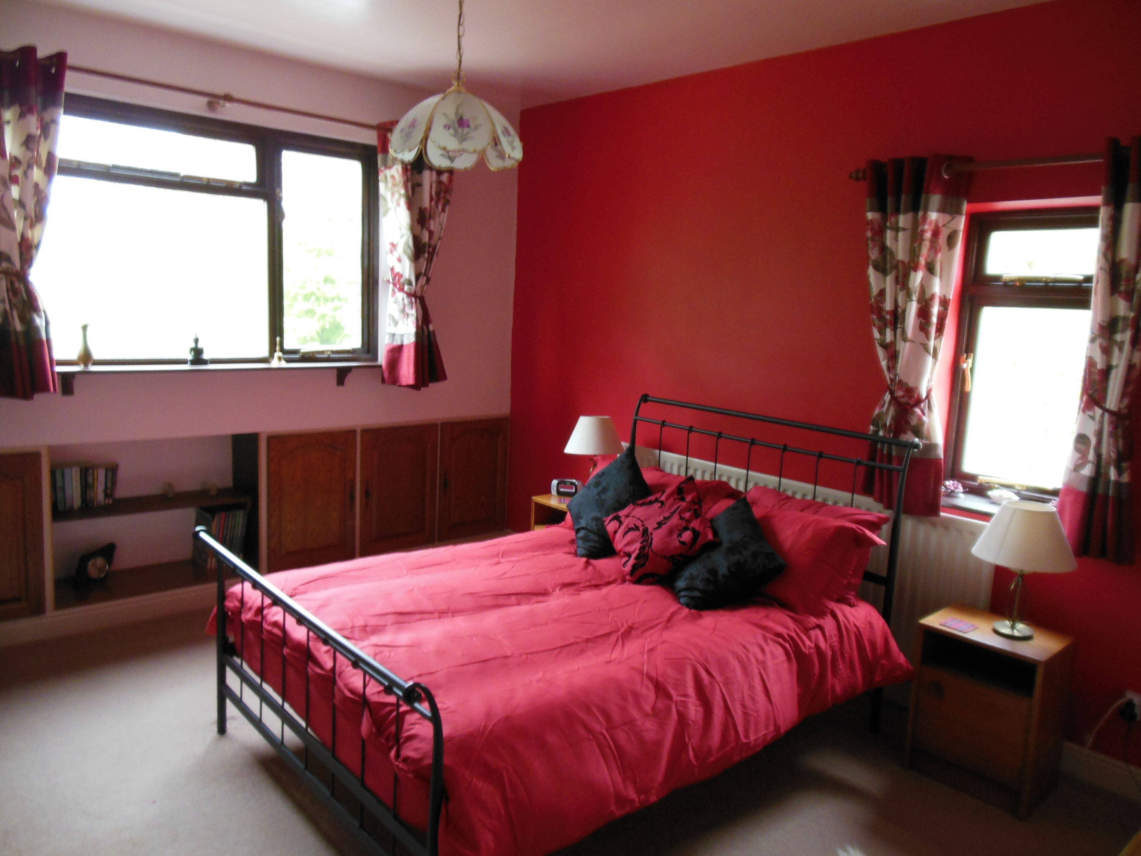 A bedroom with a red bedspread, two bedside lamps, a window with floral curtains, and a wooden wardrobe.