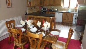Un coin repas avec des chaises en bois et une table dressée pour six personnes, une cuisine avec des meubles en bois et un réfrigérateur, et une fenêtre qui laisse entrer la lumière naturelle.