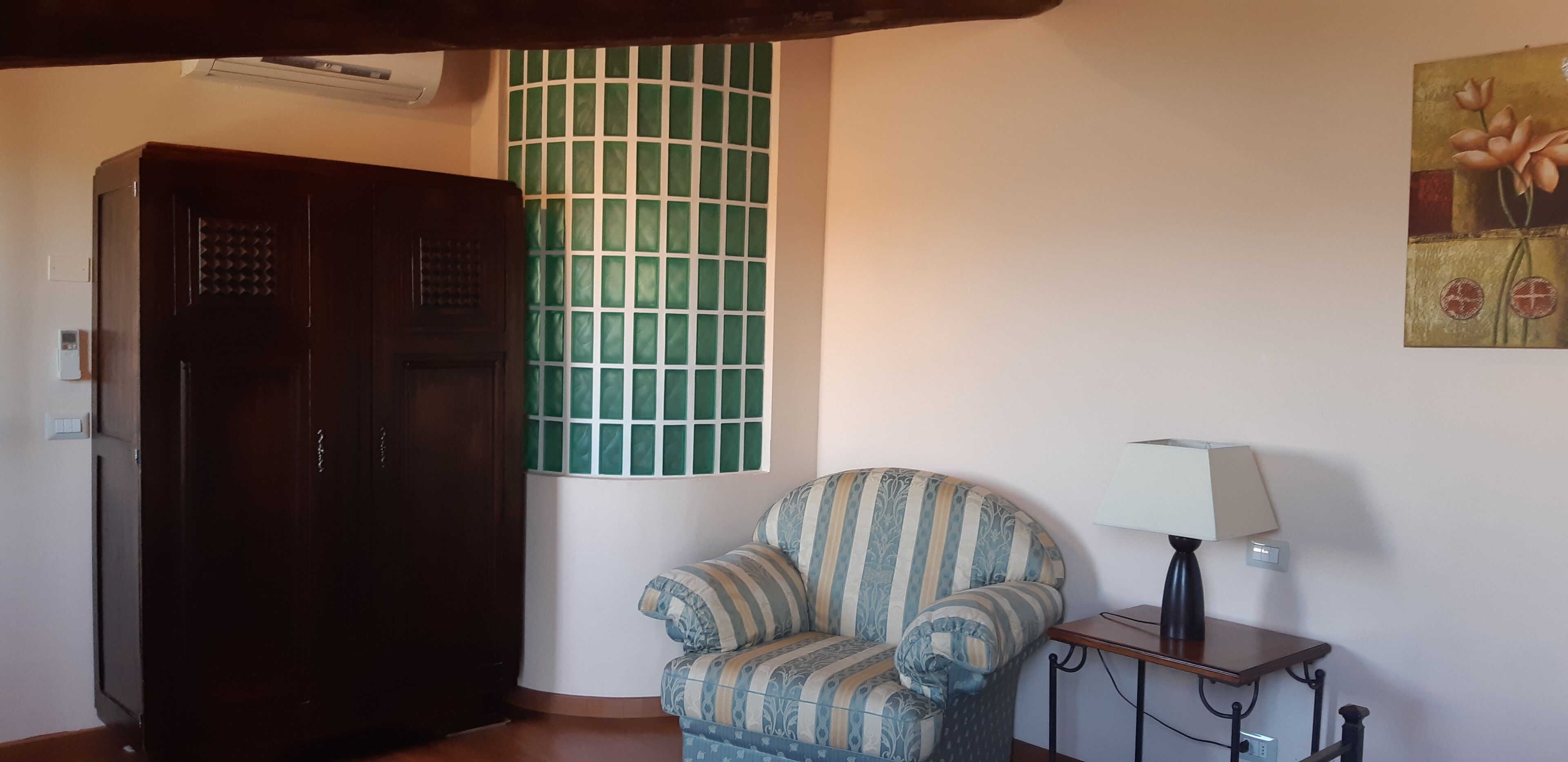 A room with a dark wooden wardrobe, a patterned armchair, a small table with a lamp, and a green-tiled window.