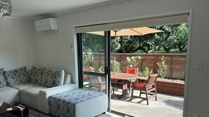 A living room with a sofa, a glass door leading to an outdoor patio with a table and chairs. - Peaceful Orchard Cottage (Omanawa)