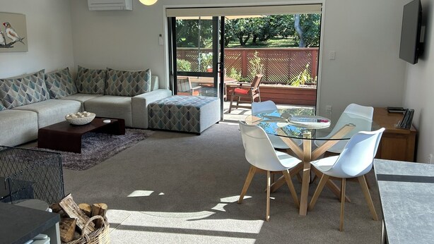 A living room with a sofa, ottoman, coffee table, and a glass dining table with chairs. There is a sliding glass door leading to an outdoor area with a patio and a view of trees. - Peaceful Orchard Cottage (Omanawa)