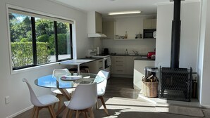 A modern kitchen with a dining area, large windows, and a wood-burning stove. - Peaceful Orchard Cottage (Omanawa)