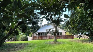 A house with a well-maintained lawn, a wooden fence, and a tree in the foreground. - Peaceful Orchard Cottage (Omanawa)